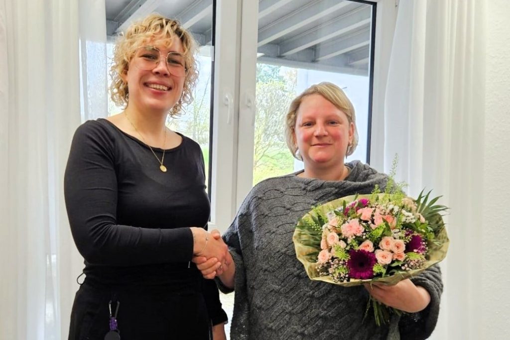 Zwei Frauen schütteln sich - lächelnd in die Kamera blickend - die Hände. Links steht die Einrichtungsleitung, rechts mit frühlingshaften Strauß mit rosa und pinken Blüten eine der beiden Jubilarinnen.