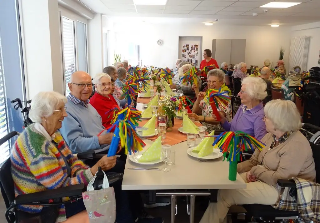 Knapp 30 ältere Menschen sitzen gut gelaunt an einer festlich gedeckten Tafel in einem großen Raum. Sie halten bunte Papierwedel in den Händen. Auf den Tischen stehen Frühlingsblumen.