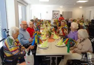 Knapp 30 ältere Menschen sitzen gut gelaunt an einer festlich gedeckten Tafel in einem großen Raum. Sie halten bunte Papierwedel in den Händen. Auf den Tischen stehen Frühlingsblumen.