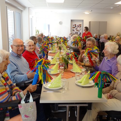 Knapp 30 &auml;ltere Menschen sitzen gut gelaunt an einer festlich gedeckten Tafel in einem gro&szlig;en Raum. Sie halten bunte Papierwedel in den H&auml;nden. Auf den Tischen stehen Fr&uuml;hlingsblumen.