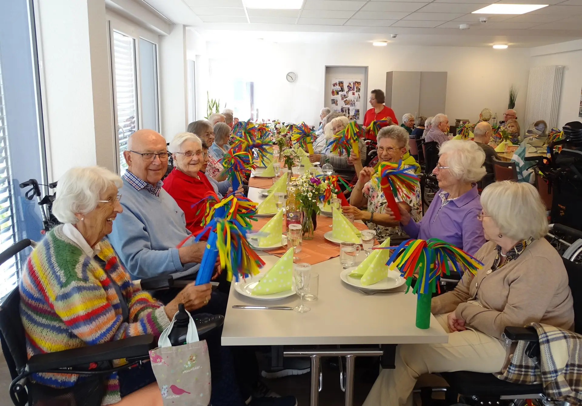 Knapp 30 ältere Menschen sitzen gut gelaunt an einer festlich gedeckten Tafel in einem großen Raum. Sie halten bunte Papierwedel in den Händen. Auf den Tischen stehen Frühlingsblumen.