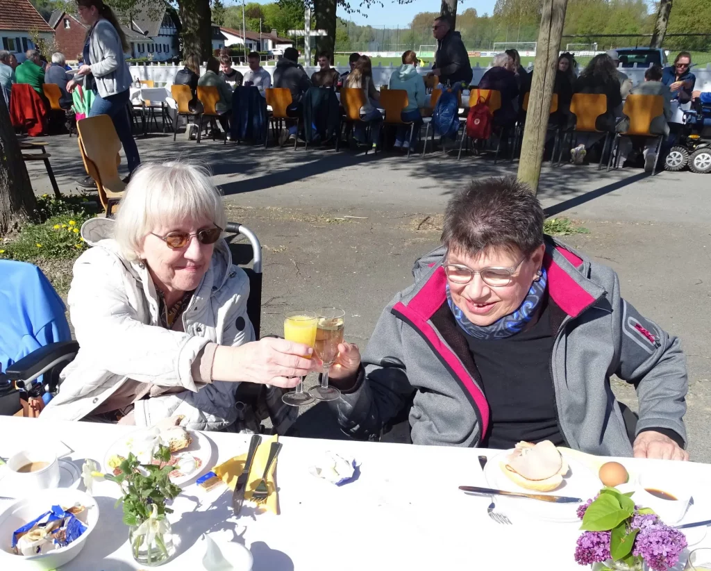 Zwei Menschen prosten sich mit Sektgläsern zu. Sie sitzen an einem gedeckten Tisch in der Sonne unter freiem Himmel. Im Hintergrund sitzen weitere Menschen an einer langen Tafel. In einem Glas ist Orangensaft, im anderen vermutlich Sekt.