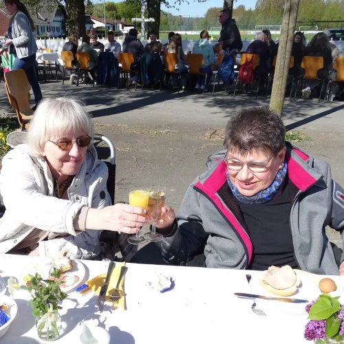 Zwei Menschen prosten sich mit Sektgl&auml;sern zu. Sie sitzen an einem gedeckten Tisch in der Sonne unter freiem Himmel. Im Hintergrund sitzen weitere Menschen an einer langen Tafel. In einem Glas ist Orangensaft, im anderen vermutlich Sekt.