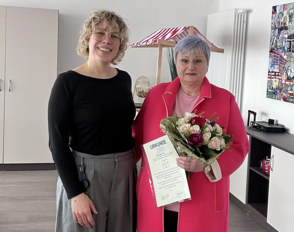 Zwei Frauen stehen lächelnd Seite an Seite: Links die Einrichtungsleitung, rechts mit üppigem Rosenstrauß mit zarten rosa und dunklen Rot-Tönen sowie einer Urkunde eine der beiden Jubilarinnen.