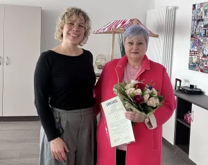 Zwei Frauen stehen lächelnd Seite an Seite: Links die Einrichtungsleitung, rechts mit üppigem Rosenstrauß mit zarten rosa und dunklen Rot-Tönen sowie einer Urkunde eine der beiden Jubilarinnen.
