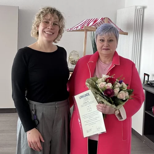 Zwei Frauen stehen l&auml;chelnd Seite an Seite: Links die Einrichtungsleitung, rechts mit &uuml;ppigem Rosenstrau&szlig; mit zarten rosa und dunklen Rot-T&ouml;nen sowie einer Urkunde eine der beiden Jubilarinnen.