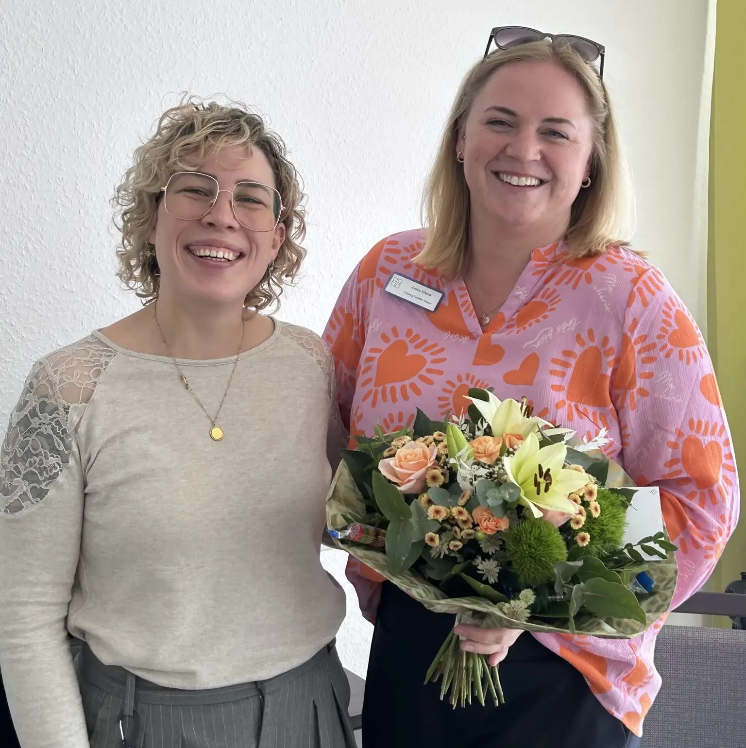 Zwei Frauen stehen lachend Arm in Arm: Links Einrichtungsleitung, rechts mit modernem Blumenstrauß mit rosa-orange-gelb-Tönen die Jubilarin.