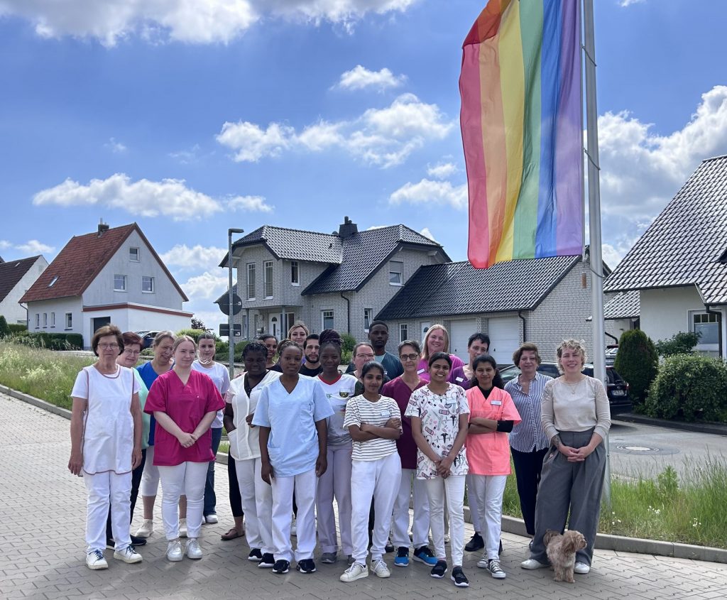 Ca. 20 Personen - Mitarbeitende des Haus Phöbe - stehen unter freiem Himmel vor der gehissten Regenbogenflagge.
