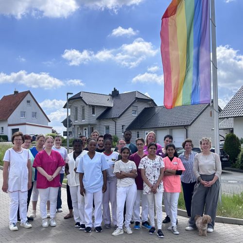 Ca. 20 Personen - Mitarbeitende des Haus Ph&ouml;be - stehen unter freiem Himmel vor der gehissten Regenbogenflagge.