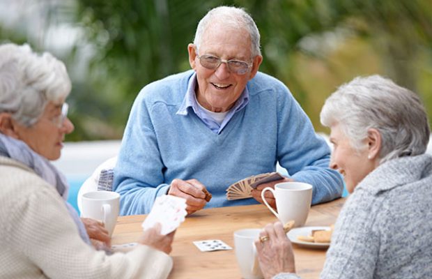 Drei ältere Menschen sitzen gesellig beisammen, trinken Kaffee und spielen Karten.